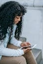 Pensive young African American woman wearing casual clothes while surfing on tablet and sitting on stone stairs near building in daylight in street in town